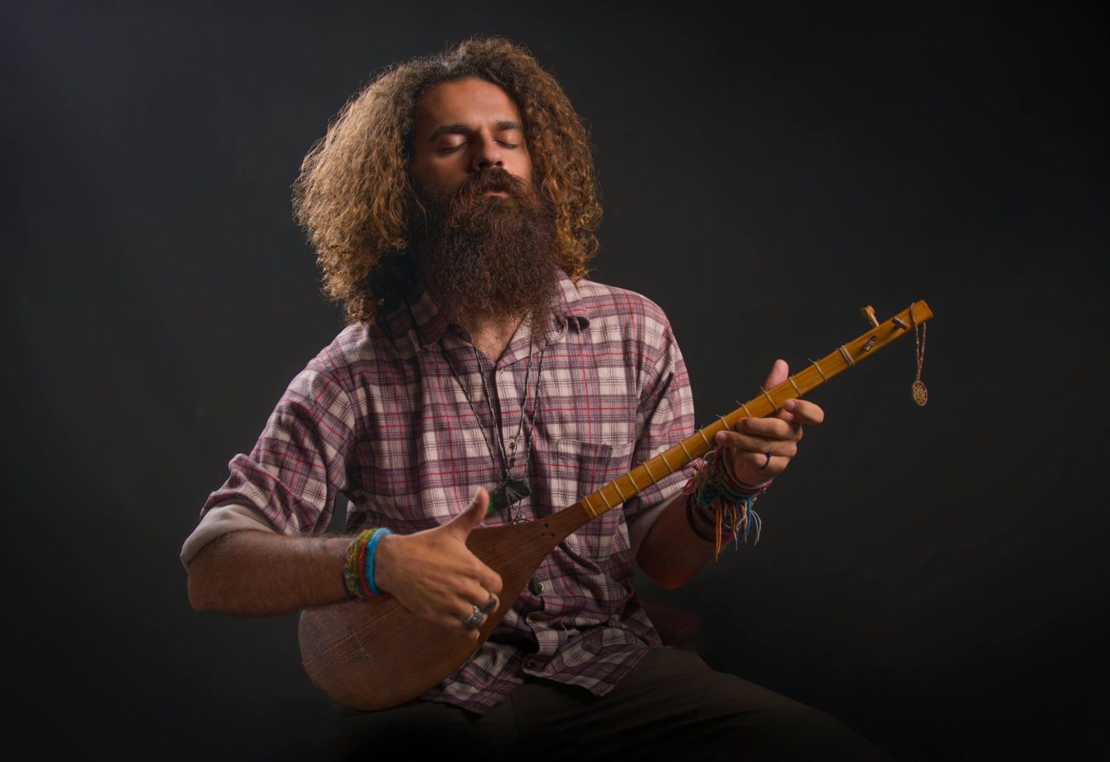 Bearded musician in a plaid shirt playing a setar against a dark background.